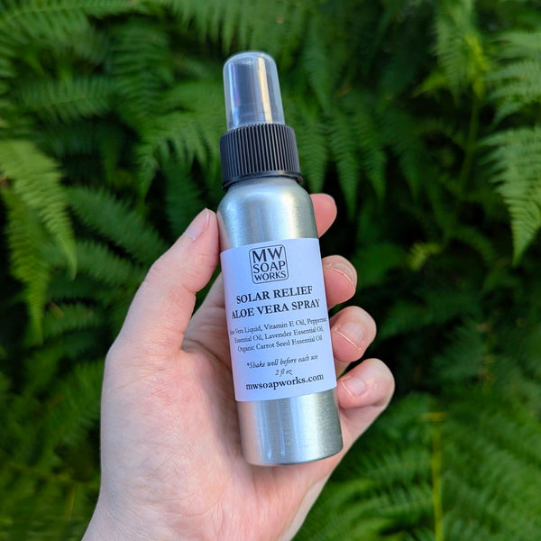 A hand holds up an aluminum bottle of MW Soapworks Solar Relief Aloe Vera Spray against a green fern background