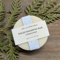 A round solid shampoo bar sits on a wooden background with a cedar branch. The label shows it is a four ounce Unscented Solid Shampoo Bar from MW Soapworks.