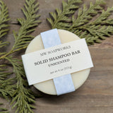 A round solid shampoo bar sits on a wooden background with a cedar branch. The label shows it is a four ounce Unscented Solid Shampoo Bar from MW Soapworks.