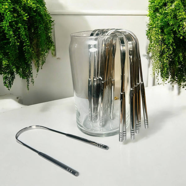A u-shaped stainless steel tongue scraper sits on a white table with plants in the background. There is a jar nearby that holds several more tongue scrapers.