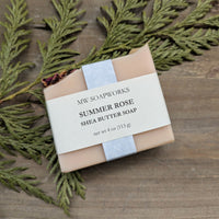 A four ounce bar of MW Soapworks Summer Rose Shea Butter Soap sits on a wooden background with cedar branches. The soap is a light pink with some rose petals along the top edge.