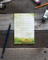 A lined notepad sits against a wooden background surrounded by art supplies. The pad features a watercolor illustration of a banana slug and the words "Take It Slow."