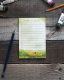 A lined notepad sits against a wooden background surrounded by art supplies. The pad features a watercolor illustration of a banana slug and the words "Take It Slow."