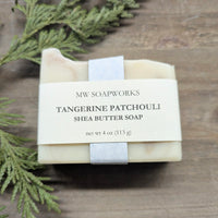 A 4 ounce bar of Tangerine Patchouli Shea Butter Soap from MW Soapworks sits on a wooden background near a cedar branch.