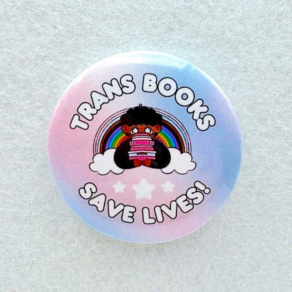 A cartoon queer cutie sits with a pile of books in front of a rainbow with the words "Trans Books Save Lives" around them on a pin.