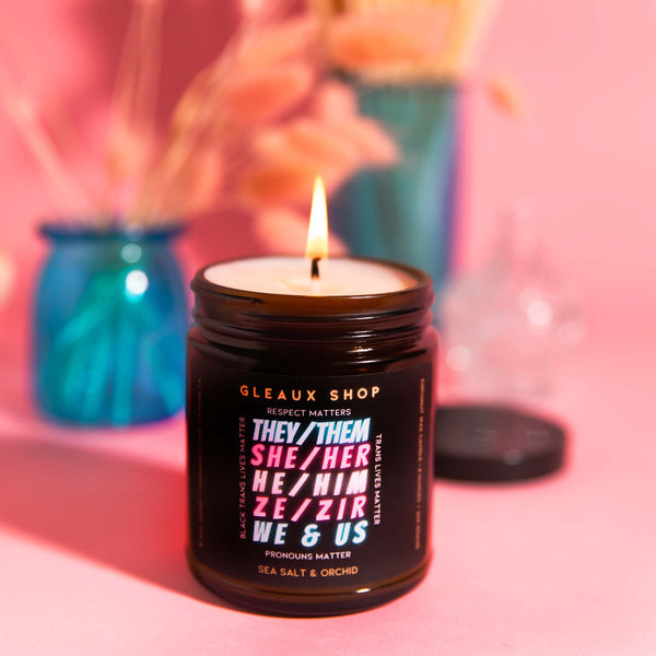 A candle in an amber jar sits on a pink table with blue accents. The candle reads "Respect Matter, Trans Lives Matter, Pronouns Matter, Black Trans Lives Matter" and lists various pronoun combinations in trans flag colors.