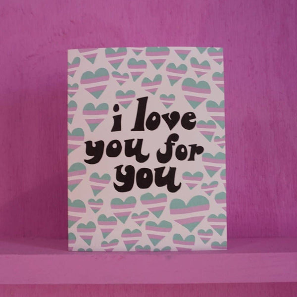 A greeting card sits against a pink background. The card is white and covered in hearts in the trans flag colors - light blue, white, and pink. In big black fun letters the card says "I love you for you."
