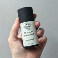 A hand holds up a black paperboard tube of MW Soapworks All Natural Lotion Stick.