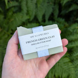 A hand holds up a bar of MW Soapworks French Green Clay Shea Butter Soap in front of PNW ferns.