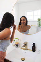 A woman stands in front of a bathroom mirror using an eco-friendly biodegradable charcoal dental floss pick