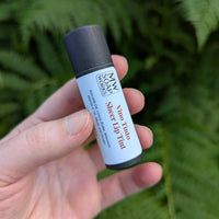 A hand holds up an ecofriendly tube of MW Soapworks Vino Tinto Sheer Lip Balm, which is a subtle red color.