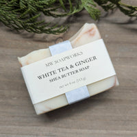 A 4 ounce bar of MW Soapworks White Tea and Ginger Shea Butter Soap sits on a wooden background near a cedar branch.