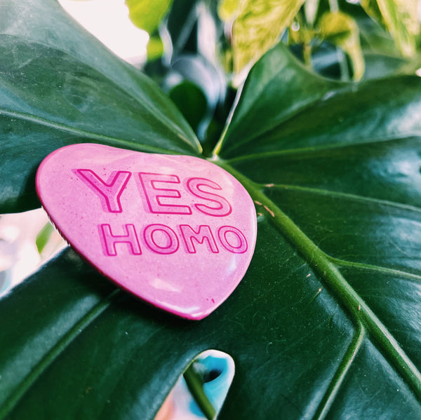 A pink heart button sits on a monstera leaf. The button says "yes homo."