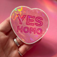 On a pink background, a hand holds a heart shaped sticker. The sticker is pink and says "yes homo."