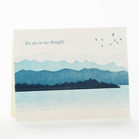 A letterpress greeting card features several layers of blue islands printed in front of mountains. Text above reads "You are in our thoughts."