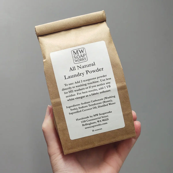 A hand holds up a 16 ounce brown paper roll-top bag of MW Soapworks All Natural Laundry Powder.