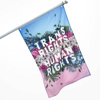 A flag featuring a floral pattern in the light blue, light pink and white of the Trans Pride flag features big block letters in white declaring Trans Rights are Human Rights.