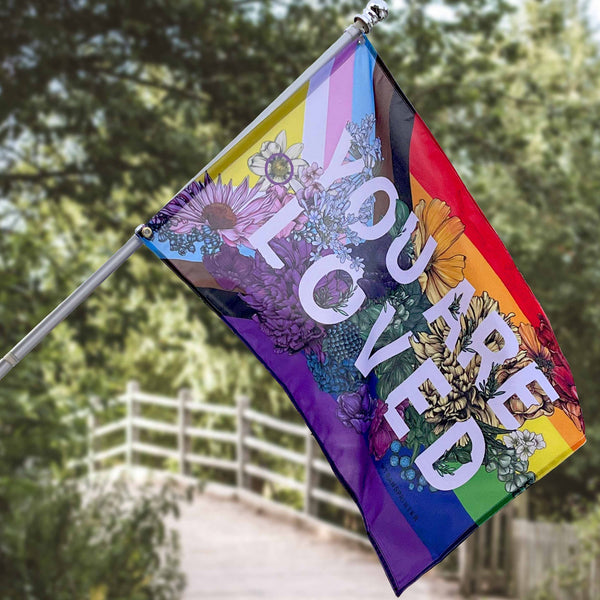 An inclusive LGBTQIA+ floral themed flag features big white block letters across it: You are loved. Flag hangs from a pole in front of a garden area.